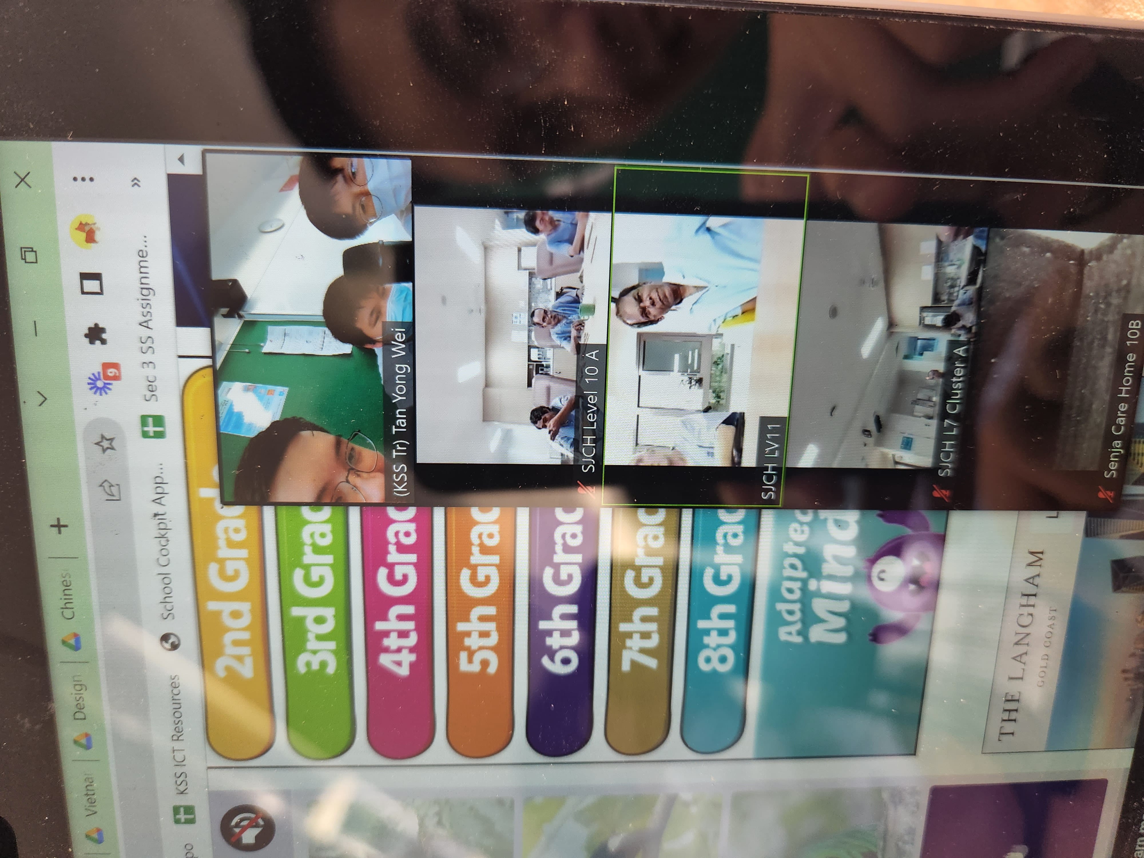 A computer screen showing a video call with multiple participants labeled SJCH levels, beside a grade-level menu website.
